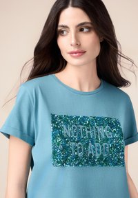 Camiseta de manga corta en color azul agua con un parche rectangular de lentejuelas que presenta el texto "NADA QUE AÑADIR" en un color azul contrastante. Textura de tela suave.