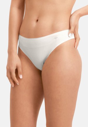 Nainen, joka pukeutuu saumattomaan valkoiseen bikinityyppiseen alusvaatteeseen, seisoo neutraalia taustaa vasten, toinen käsi levollisesti lanteillaan.