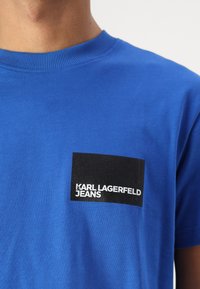 Mėlynas medvilninis marškinėliai su banguota iškirpte ir juodu stačiu kampu užklijuota etikete, kurioje baltais rašmenimis parašyta "KARL LAGERFELD JEANS".