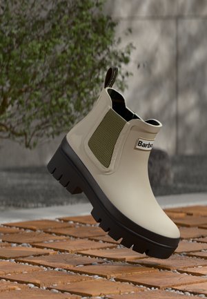 Beige rubber Chelsea-laarzen met groene elastische zijpanelen en een zwarte profielzool, voorzien van een logo aan de zijkant en een trekband aan de achterkant.