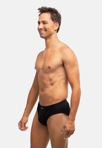 Slip in cotone nero con una vestibilità aderente, caratterizzato da una texture liscia e una cintura elastica con logo. Indossato da un modello maschile di profilo.