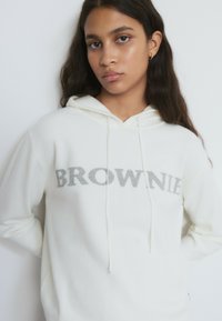 Sudadera con capucha blanca con la palabra "BROWNIE" impresa en gris. Confeccionada con tela suave, cuenta con capucha con cordón y un ajuste holgado. La textura parece lisa.