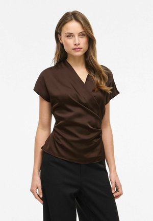 Kvinde iført en mørkebrun, kortærmet slå-om-bluse med rynkede sider og sorte bukser, stående foran en ensfarvet hvid baggrund.