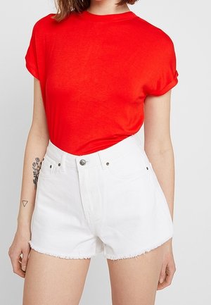 Camiseta roja de manga corta combinada con pantalones cortos de mezclilla blancos con dobladillo deshilachado y cierre frontal de botones. Piel visible con un tatuaje en el brazo.