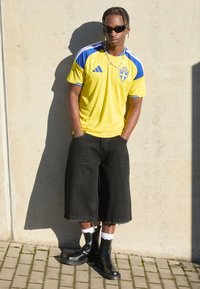 Κίτρινη φανέλα Adidas με μπλε λεπτομέρειες και σουηδικό έμβλημα, μαύρες φαρδιές βερμούδες, μαύρα μποτάκια αστραγάλου και λευκές κάλτσες. Απαλή υφή.