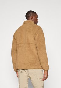 Beige fleecejack met hoge kraag, gestructureerde buitenkant en lange mouwen, gecombineerd met lichte cargobroek.