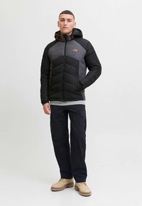 Svart och grå vadderad jacka med huva, med dragkedjeförslutning framtill, orange accenter och texturerade paneler. Bärs med cargobyxor och stövlar.