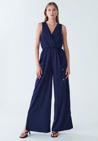 Temno modra kombinezonka z ovijalnim dizajnom, brez rokavov, z nastavljivo vrvico v pasu in širokimi hlačnicami s stranskimi razporki. Gladek tekstura tkanine.