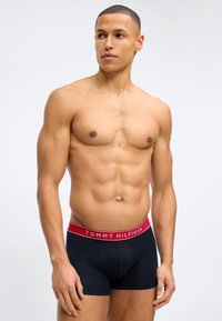 Μυώδης άνδρας που στέκεται χωρίς πουκάμισο, φορώντας ναυτικό μπλε boxer Tommy Hilfiger με κόκκινη μέση, κοιτάζοντας προς τα αριστερά σε απλό φόντο.