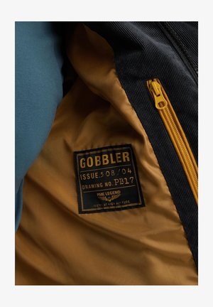 Wnętrze kurtki z czarnym nadrukiem z materiału kordurowego, żółtą podszewką oraz metką z napisem "GOBBLER" i specyfikacjami w wyboldowanej czcionce. Zamek w kolorze złotym.