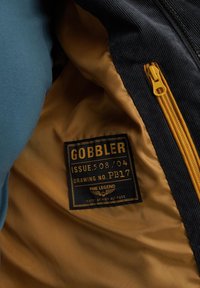 Interior de una chaqueta de marina con forro amarillo y una etiqueta negra. La etiqueta presenta texto dorado con detalles del producto, y la textura del tejido parece suave.