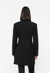 Svart skreddersydd blazer med lange ermer, strukturerte skuldre og innsnevret midje. Har knappdetaljer på ermene og en glatt tekstur.