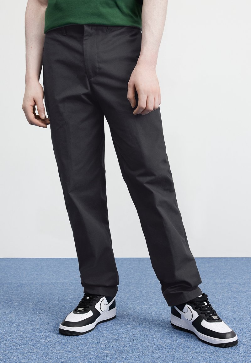 Black trousers with a smooth texture, straight leg design, and a mid-rise waist, paired with black and white sneakers.