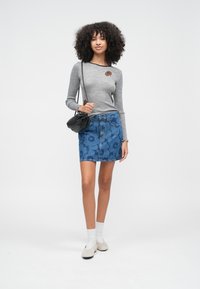 Göndör hajú nő szürke hosszú ujjú felsőt, kék virágos farmer szoknyát, fehér zoknit, bézs cipőt és fekete válltáskát visel.