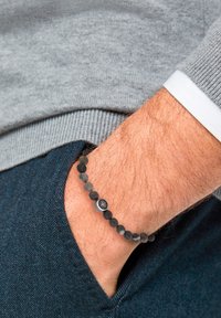 Bracciale nero di perline con pietre lisce e texture, caratterizzato da un accento metallico. Indossato su un polso con capelli chiari, su sfondo di tessuto grigio.