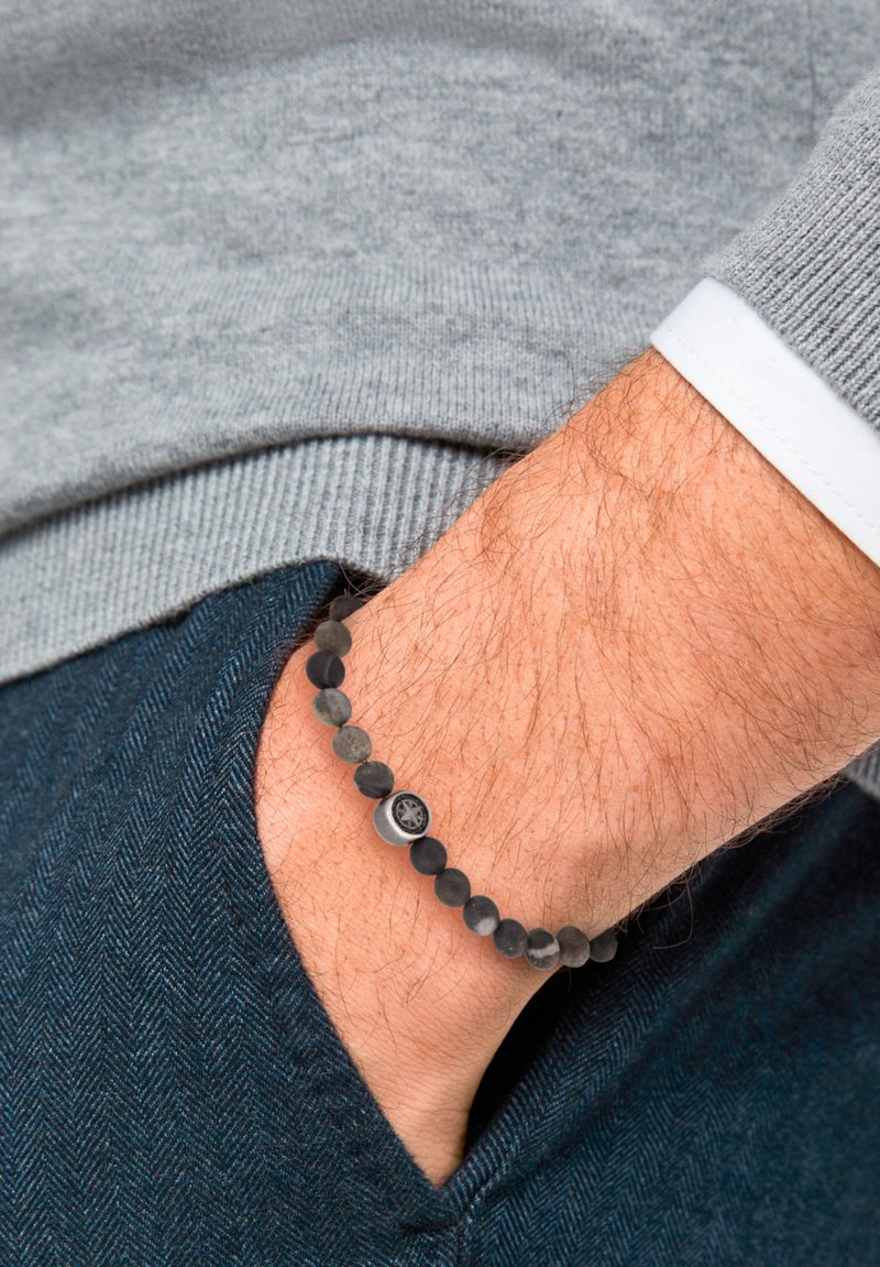 Bracciale nero di perline con pietre lisce e texture, caratterizzato da un accento metallico. Indossato su un polso con capelli chiari, su sfondo di tessuto grigio.