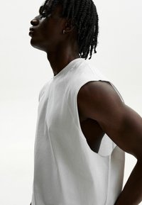 Hvid ærmeløst skjorte med crew neck og udskårne detaljer i siden, lavet af blødt stof med en glat tekstur. Simpelt design, minimale accenter.
