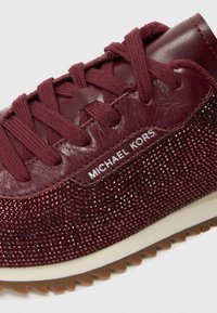 Bordowe trampki z teksturowymi i błyszczącymi panelami, dopasowanymi sznurowadłami z materiału, białym logo "MICHAEL KORS" oraz brązową, żłobioną podeszwą.