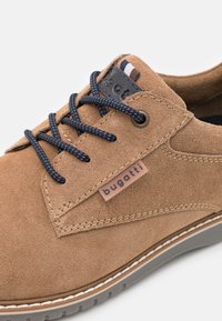 Zapato de ante marrón claro con cordones azul marino, que presenta un parche con logo y costuras en contraste a lo largo de la suela blanca.