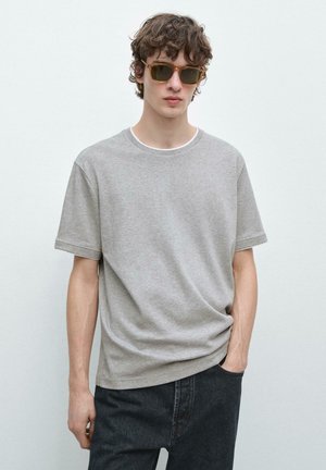 Giovane con capelli ricci che indossa occhiali da sole marroni, una t-shirt grigio chiaro a maniche corte e jeans scuri, in piedi davanti a uno sfondo bianco uniforme.