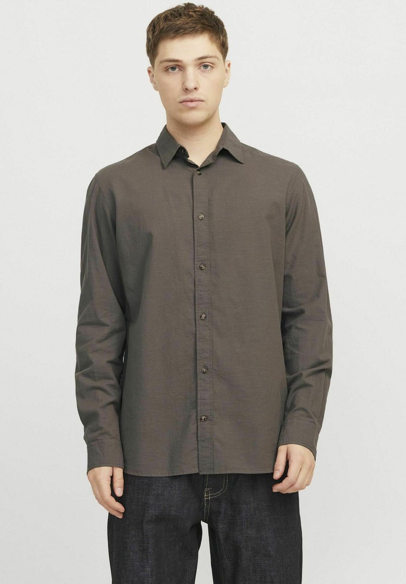 Camisa de manga larga marrón, abotonada, en una tela ligera con cuello, botones frontales y un corte relajado, combinada con pantalones vaqueros oscuros.