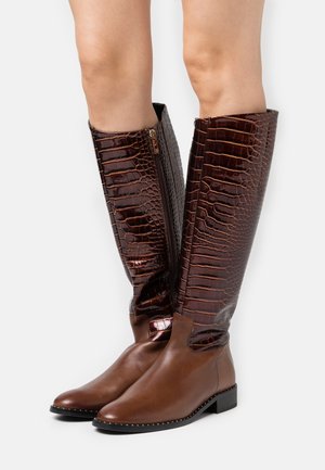 Bottes montantes en cuir marron avec une tige embossée crocodile, une fermeture éclair latérale, et une semelle plate contrastante ornée de clous métalliques le long du bord.