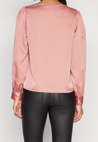 Blouse en satin rose clair à manches longues, épaules froncées et poignets boutonnés, vue de dos, associée à un pantalon noir ajusté.