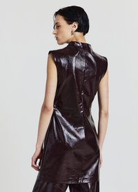 Braunes ärmelloses Kleid aus Vinyl mit hohem Kragen, das eine zentrale Rückennaht und seitliche Schlitze für Bewegungsfreiheit aufweist. Glatte, glänzende Textur.