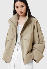 Giacca oversize color khaki realizzata in tessuto leggero, con due ampie tasche anteriori, colletto alto e dettagli con zip.