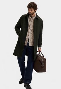 Donkergroene wollen jas, beige overhemd met visgraatpatroon, donkerblauwe jeans, zwarte schoenen, draagt een bruine leren weekendtas. Eenvoudig, gestructureerd ontwerp.