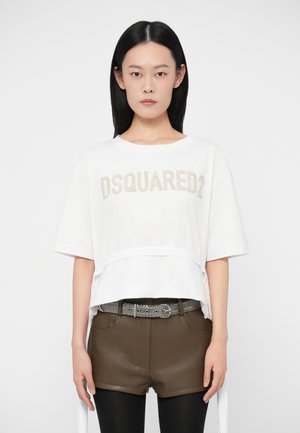 Žena stojí čelom dopredu, oblečená v bielom tričku DSQUARED2, hnedých kožených šortkách s pások s vzorom a čiernych legínach.