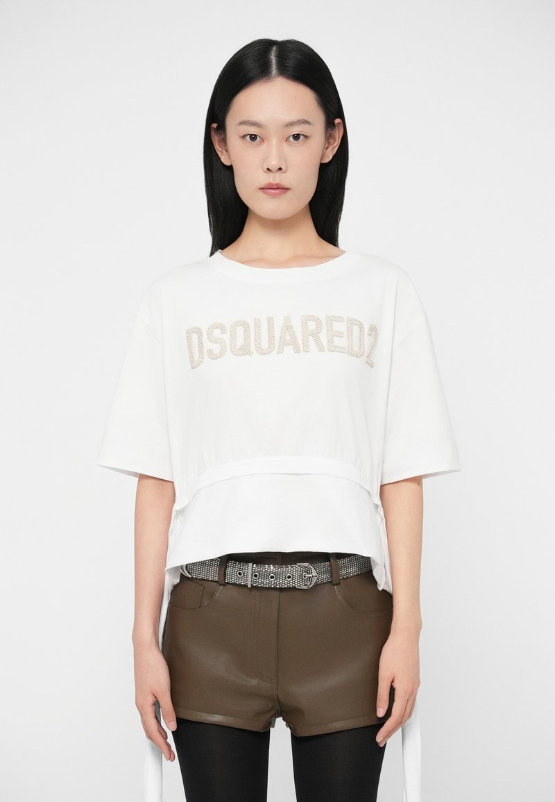 Žena stojí čelom dopredu, oblečená v bielom tričku DSQUARED2, hnedých kožených šortkách s pások s vzorom a čiernych legínach.
