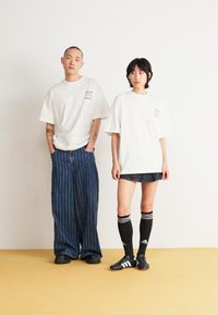 Two models stand side by side. The male model wears a loose white t-shirt and striped pants. The female model wears a white t-shirt with a denim skirt and black knee-high socks.