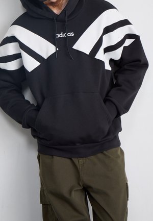 Personne portant un sweat à capuche noir avec des rayures diagonales blanches et un logo, associé à un pantalon cargo vert olive, mains dans la poche du sweat.