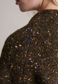 Brązowy sweter o fakturze z kolorowymi kropkami w odcieniach różu, niebieskiego i żółtego rozmieszczonymi na całej tkaninie. W widocznym na zdjęciu okrągłym dekolcie.