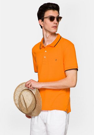Pomarańczowa koszulka polo z czarnymi wykończeniami, krótkie rękawy i logo na piersi. Model trzyma słomkowy kapelusz, nosi okulary przeciwsłoneczne. Neutralne tło.