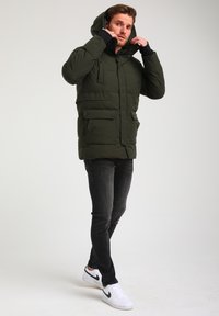 Giacca imbottita verde scuro con cappuccio, dotata di grandi tasche frontali, chiusura con zip e polsini a coste. Indossata con jeans neri e sneakers bianche.