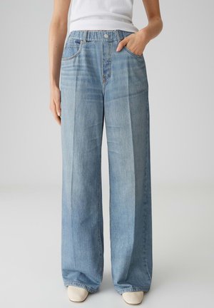 Persoon draagt lichtblauwe wijd uitlopende jeans met voorplooien, één hand in de zak, gecombineerd met witte schoenen en een wit mouwloos topje.