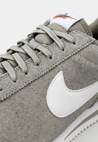 Sivé semišové tenisky s bielym Nike swooshom, textúrovaným vrchom, plochými sivými šnúrkami a bielou podrážkou. Obsahujú prešitý šev pozdĺž okraja.