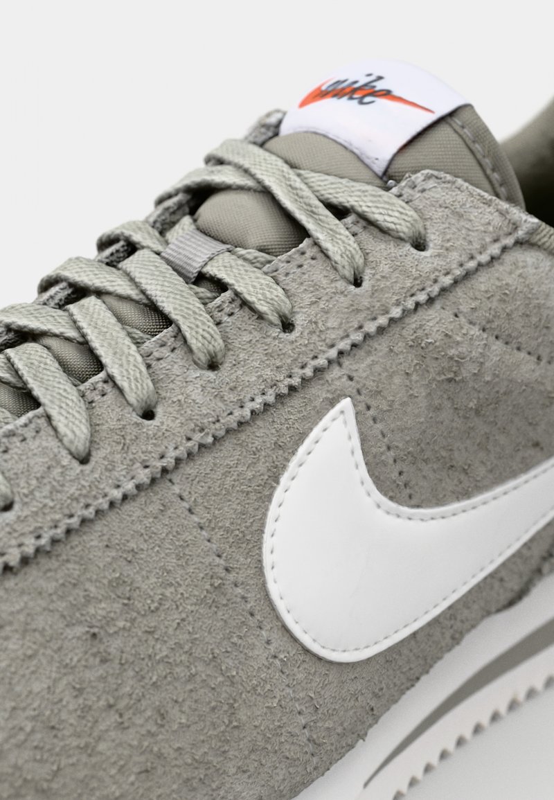 Sneaker in suede grigio con il logo bianco Nike, tomaia texture, lacci piatti grigi e suola bianca. Presenta una cucitura lungo il bordo.