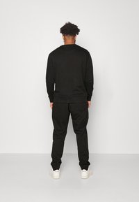 Zwarte sweatshirt en joggers, gemaakt van zachte stof. Kenmerken zijn ribbels aan de mouwen en tailleband, met een achterzak op de joggers. Het schoeisel is wit.