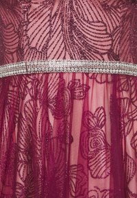 Burgundowa sukienka z błyszczącą koronką w kwiatowe wzory i przezroczystymi warstwami, z metalicznym paskiem ozdobionym srebrnymi cyrkoniami podkreślającym talię.