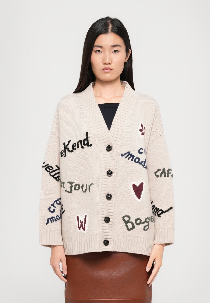 Cardigan lavorato a maglia beige con scollatura a V profonda, grandi bottoni neri e frasi ricamate in vari colori. Presenta maniche larghe e polsini a coste.