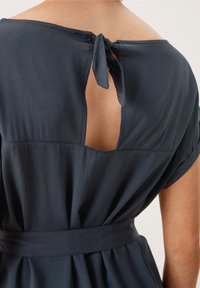Mörkblå klänning med texturerad tyg, med en nyckelhålsöppning bak, detalj med knytning i halsen och midja med bälte.