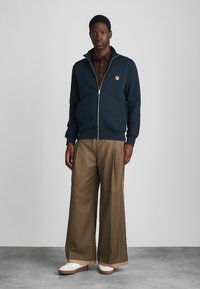 Hanorac bleumarin cu fermoar, cu guler înalt, asortat cu pantaloni largi maro. Are un mic logo portocaliu pe piept. Adidași albi.