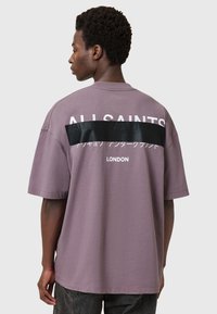 Camiseta oversized de color lavanda con un panel negro que presenta texto blanco en la parte posterior. Diseño de mangas cortas y cuello redondo.