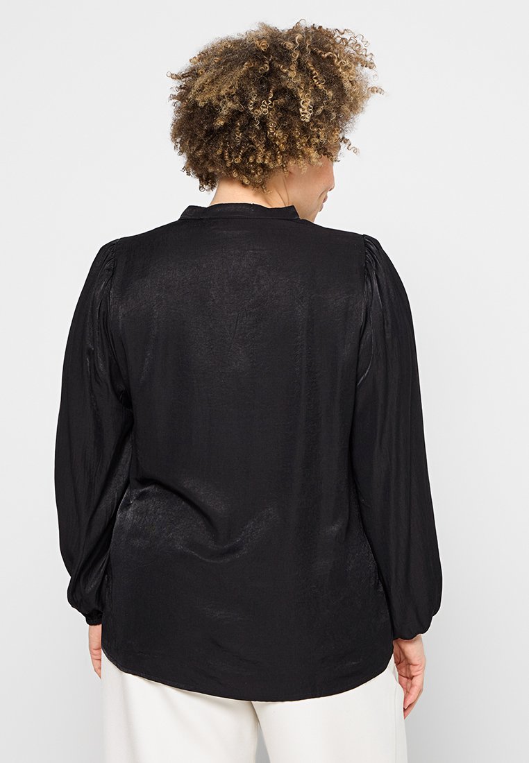 Blouse noire à manches longues en tissu lisse et texturé. Présente des épaules bouffantes et un dos boutonné avec un col simple.
