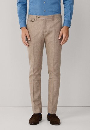 Mężczyzna ubrany w beżowe, dopasowane spodnie, niebieską koszulę na guziki oraz ciemnobrązowe mokasyny zamszowe, stojący na jednolitym tle.