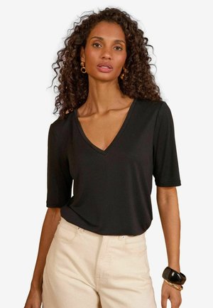 Žena s kučeravými vlasmi, oblečená v čiernom tričku s V-výstrihom a krátkymi rukávmi, béžových nohaviciach, zlatých kruhových náušniciach a čiernych a zlatých náramkoch.