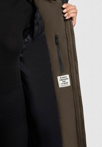 Olīvkoka zaļš svečturis ar rāvējslēdzēja kabatu kreisajā iekšpusē, ar melnu oderi. Uz birkas rakstīts: "Siltas apskāvienas no auduma."